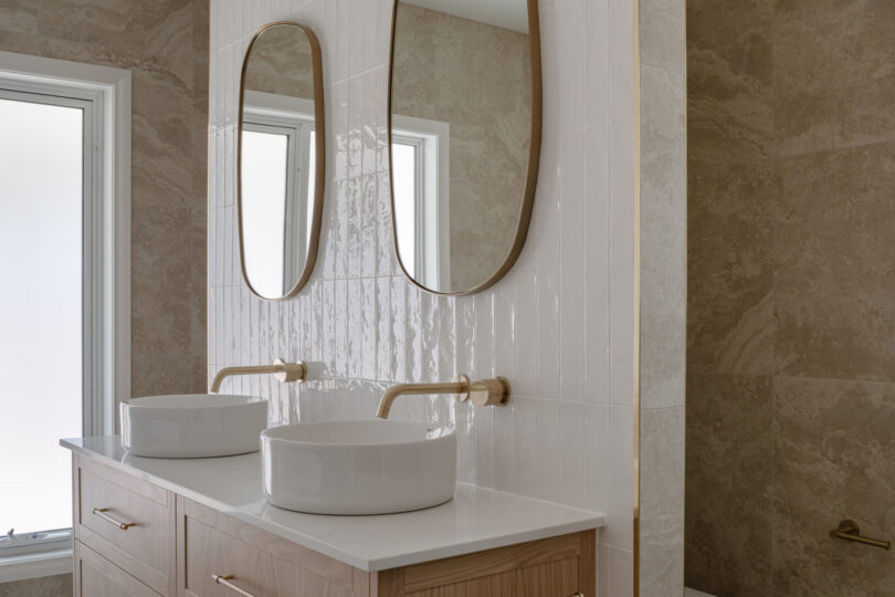 Floating timber vanity with two round vessel basins, slim oval mirrors and brushed-brass wall taps, set against vertical white feature tiles and warm travertine-look walls and floor; toilet cubicle just beyond.