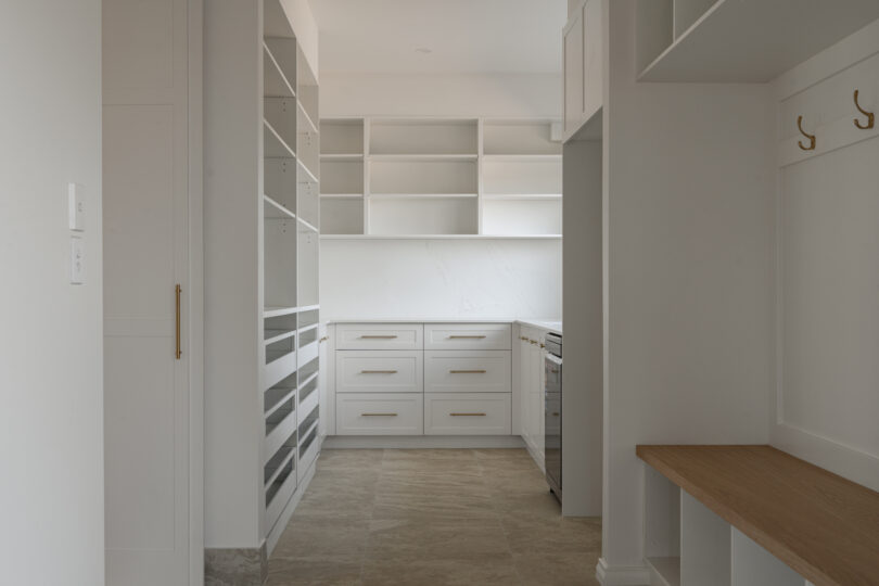 Walk-through butler’s pantry showcasing white shaker drawers, marble-look quartz splashback and benchtops, open shelving and brass hardware, all grounded by neutral stone floor tiles.