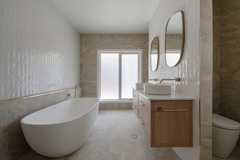 Serene main bathroom with a freestanding white oval bath beneath a frosted window, oak floating vanity with twin basins, vertical white tiles, brushed-brass fixtures and sandy stone tiles throughout.