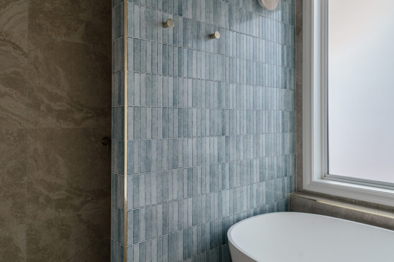 Detail of shower feature wall clad in narrow teal glazed finger tiles with two round stone shelves and brass robe hooks, contrasted by adjacent travertine-look tiles and the edge of the bath.