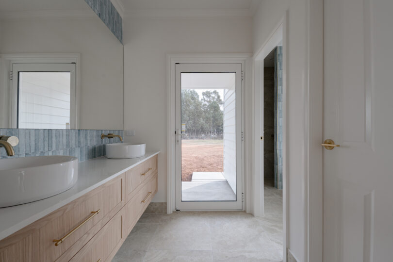 Ensuite vanity run: timber drawers, twin countertop basins and brass wall-mounted taps beneath a full-width mirror bordered by a blue tile band, with a glazed door opening to the outdoors on the right.
