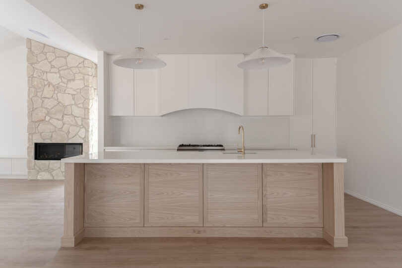 Clean, minimalist kitchen starring a light-oak island with crisp white quartz benchtop, arched white range-hood surround, brass gooseneck tap, twin pleated pendant lights and a rustic limestone fireplace column.