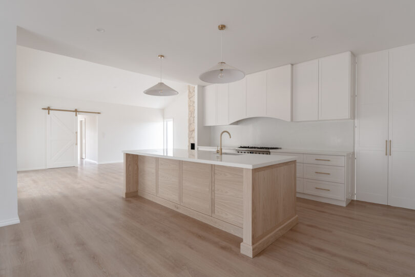 Alternate angle of the open-plan kitchen highlighting the long oak island, expansive timber flooring, white shaker cabinetry and a white barn door on a brass track beneath a gently vaulted ceiling.