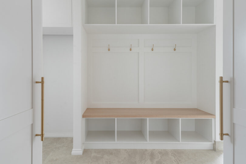 Custom mudroom joinery: white cubbies below and above an oak bench seat, framed panelling with four brushed-brass coat hooks, flanked by tall cabinetry with matching brass pulls.