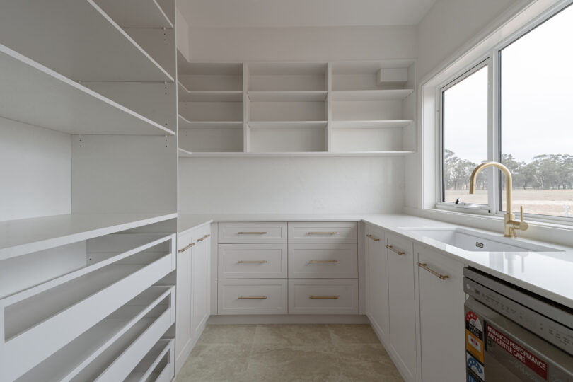U-shaped scullery with white shaker drawers, abundant open shelving, marble-look quartz surfaces, a picture window over a farmhouse sink fitted with a brass mixer tap and pull-out pantry racks along one wall.