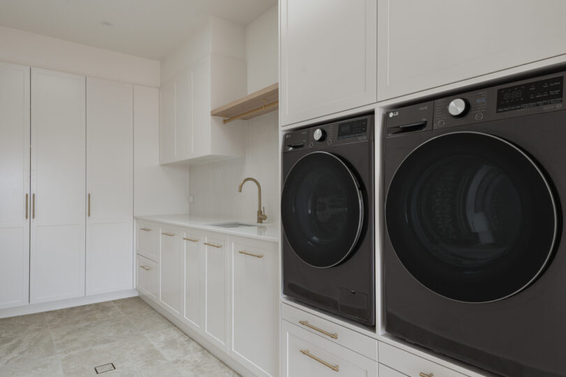 Light-filled modern-farmhouse laundry featuring white shaker cabinetry, stacked black front-loading washer and dryer, brass tapware and pulls, large travertine-look floor tiles and a square skylight overhead.