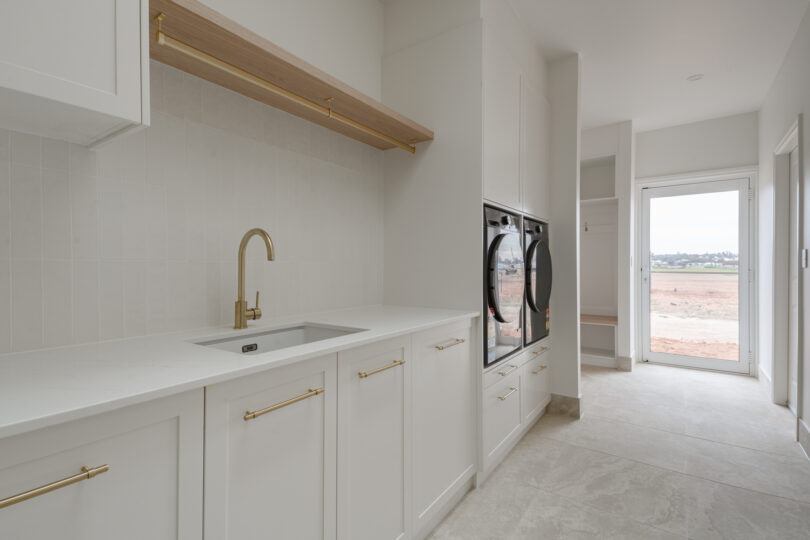 Streamlined utility-mudroom corridor with white shaker cabinets, brass pulls, brass gooseneck tap over an under-mounted sink, vertical white tiles, timber shelf and hanging rail, stacked black washer-dryer, bench nook and a glazed door opening to rural views across travertine-look floor tiles.