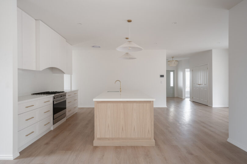 Light, open kitchen showcasing a timber-veneer island topped in white quartz, arched white range-hood surround, shaker drawers with brass hardware, stainless dual-fuel cooker, twin pleated pendants and pale timber flooring flowing into the adjoining hall.
