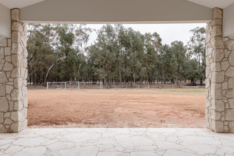 Framed outlook from the covered verandah—flagstone paving and limestone piers opening to a bare earth yard, rural gate and a backdrop of mature eucalypt woodland.