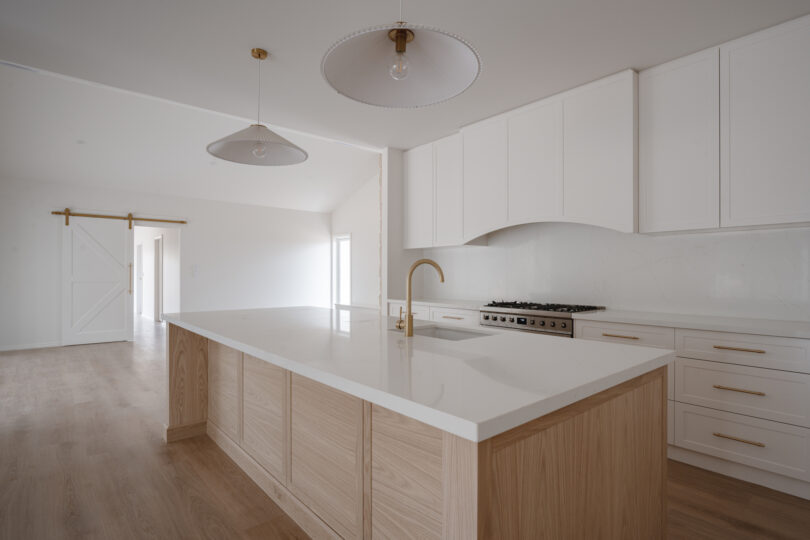 Close-angle kitchen perspective highlighting the expansive quartz island, sculptural brass mixer, pleated pendants and a white barn door on a brass track against the warm oak floor.
