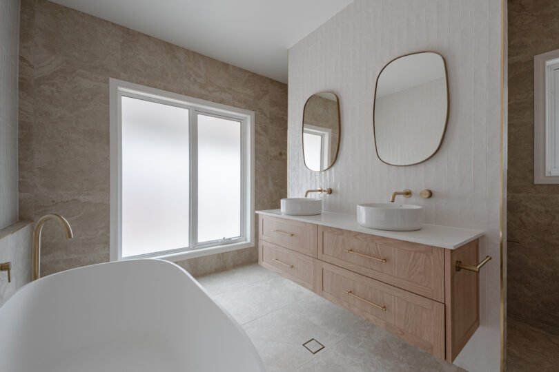 Calm main bathroom scene with a white freestanding bath beside a frosted picture window, oak floating vanity fitted with twin round basins, organic brass-framed mirrors, vertical white tiles and travertine-look walls and floor.