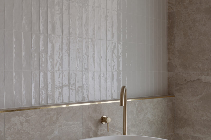 Detail of the bath zone—matte white tub, slim brass spout, glossy vertical white tiles above travertine-look slabs, finished with a sleek brass shadow-line trim.