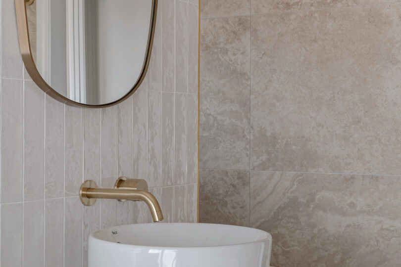 Vanity close-up: round ceramic basin, brushed-brass wall tap, slim brass-edged oval mirror and oak drawer front with brass pull against a backdrop of white vertical tiles and travertine-look stone.