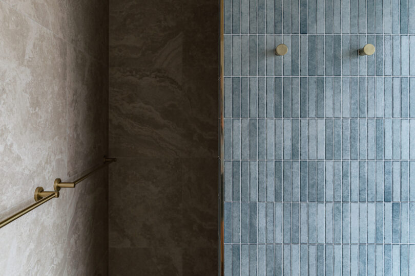 Textural shower detail showing slender teal finger tiles with twin brass robe hooks alongside a travertine-look wall fitted with a matching brass towel rail.
