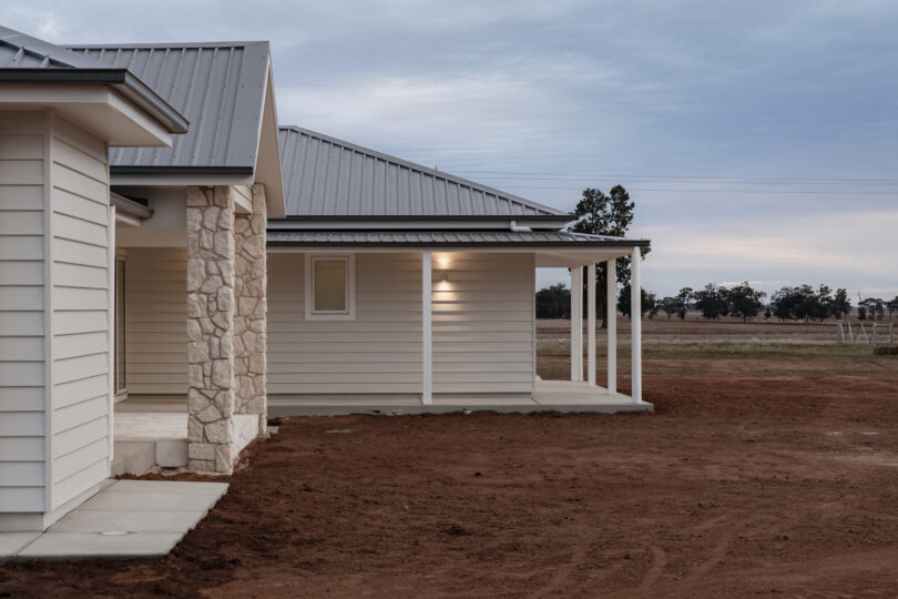 Evening side elevation of the weatherboard home showing light-grey cladding, a limestone columned veranda, standing-seam Colorbond roof and raw earth surrounds under a cloudy sky.