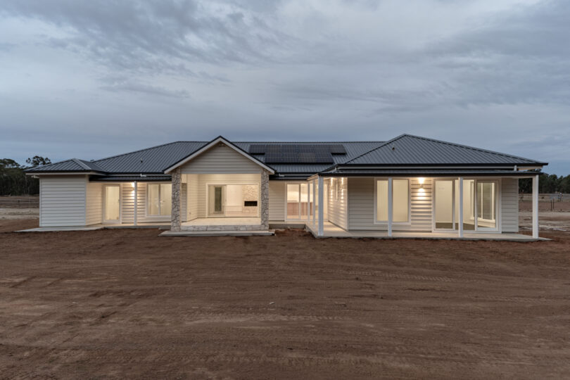 Wide rear panorama of the L-shaped residence capturing the central gabled terrace, continuous wrap-around verandas, rooftop solar array and large glazed openings glowing at twilight.