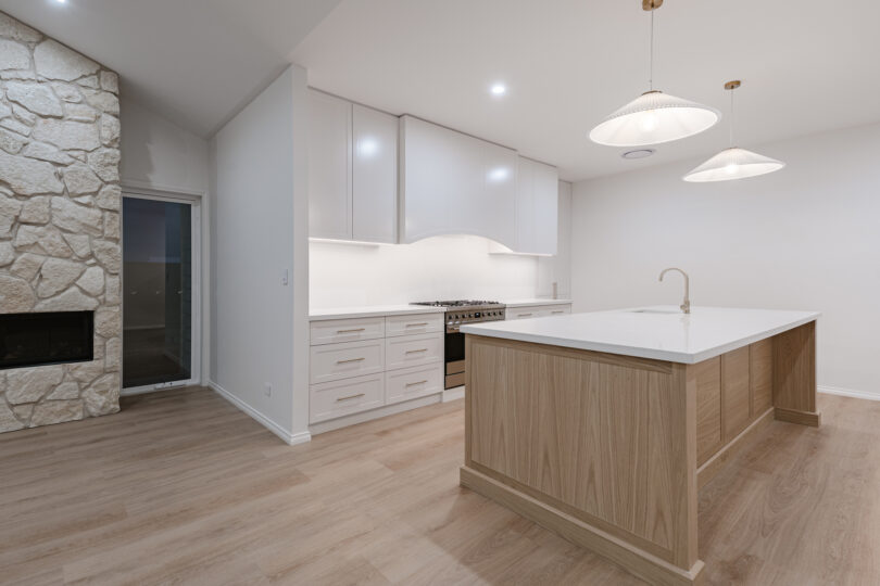 Night-time interior of the kitchen showcasing a white-quartz island with oak panelling, shaker cabinetry, stainless dual-fuel cooker, pleated pendant lights, pale oak flooring and a textured limestone fireplace wall.